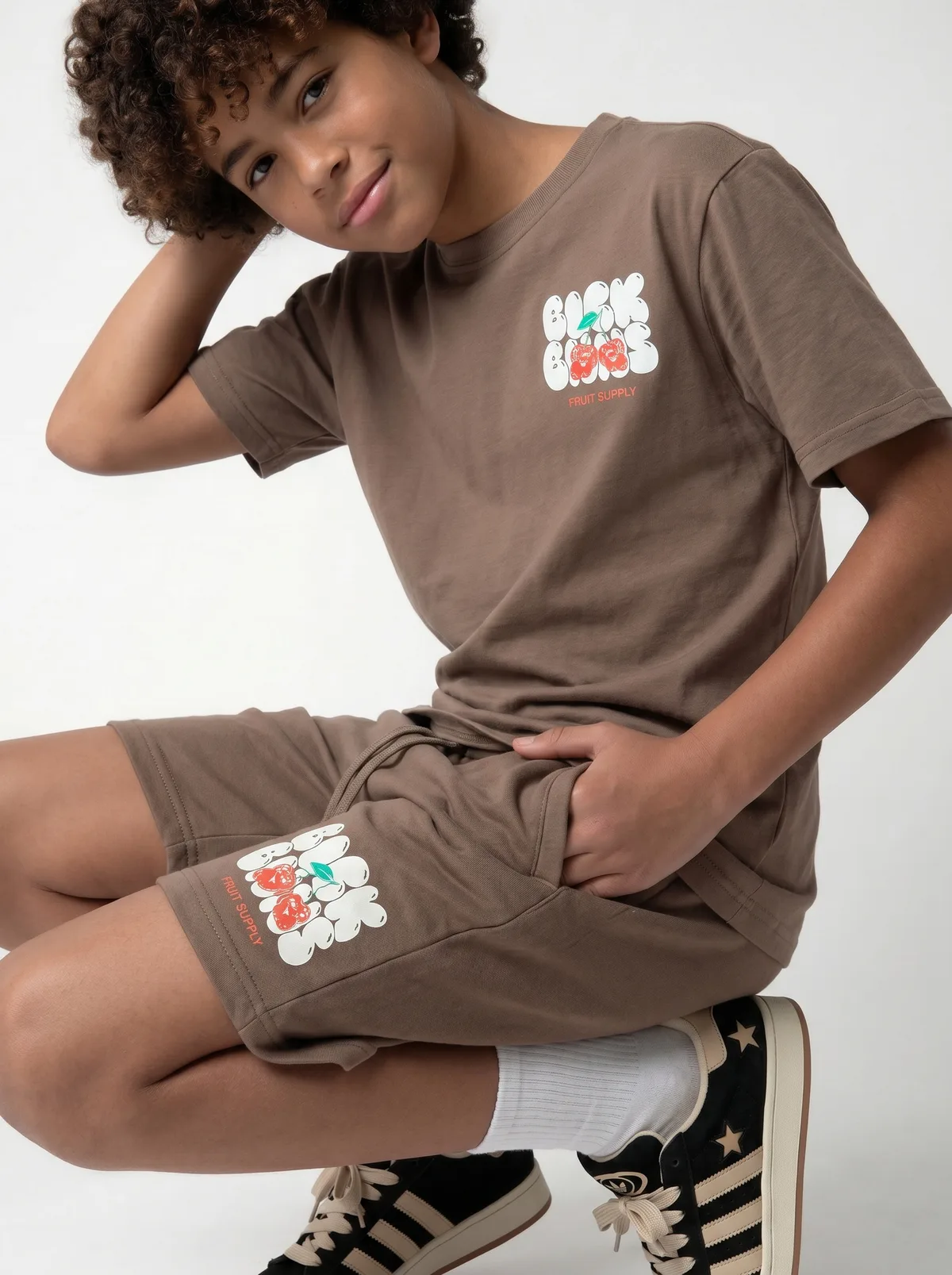 Black Bananas warm brown Fruit Supply tee chest-print detail crop showing bubble-type fruit-supply graphic with friendly orange characters on AI young model