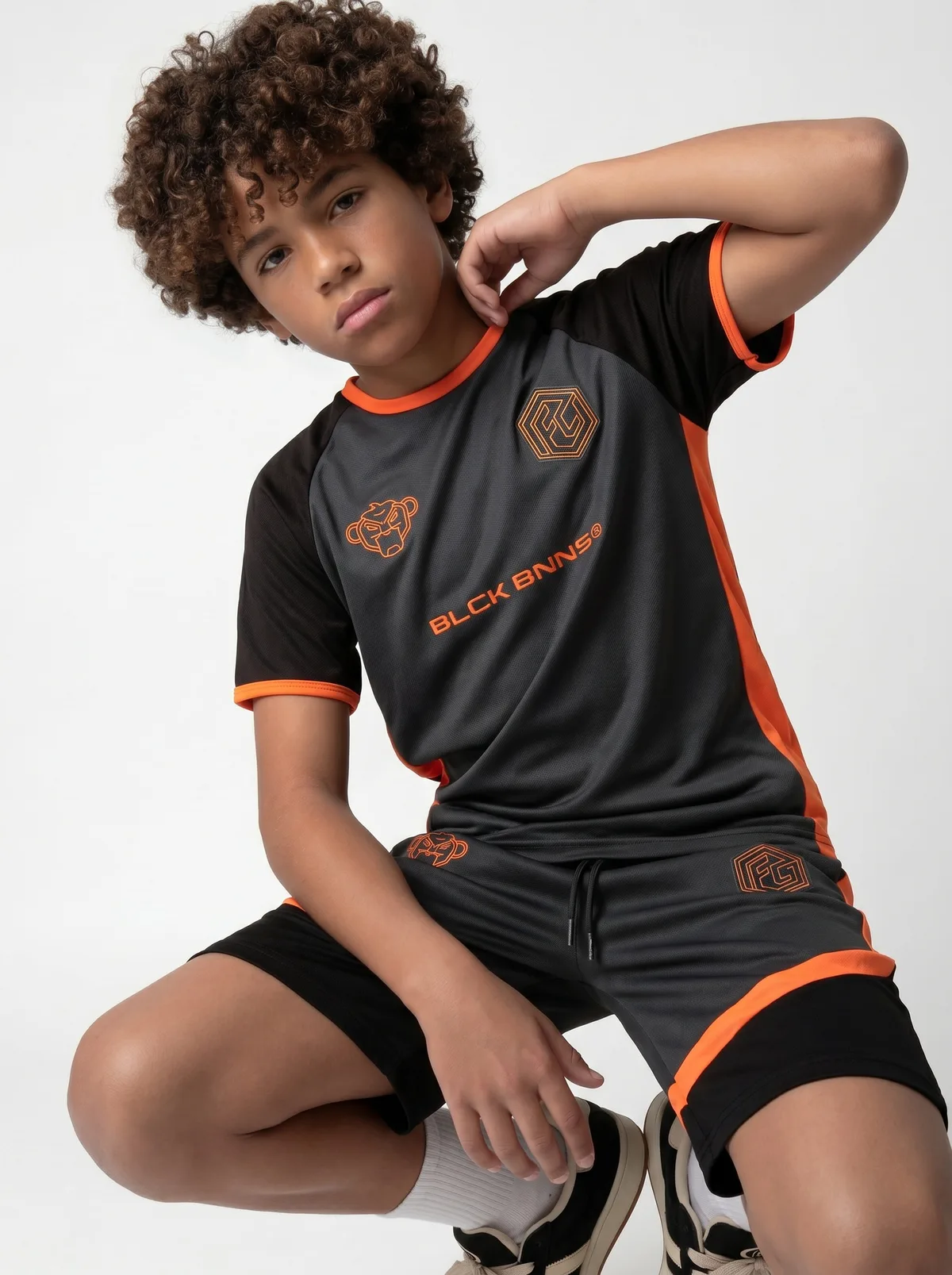 Black Bananas FC-style orange-and-charcoal football jersey three-quarter crouching editorial pose on AI young model with hand to head in clean white studio