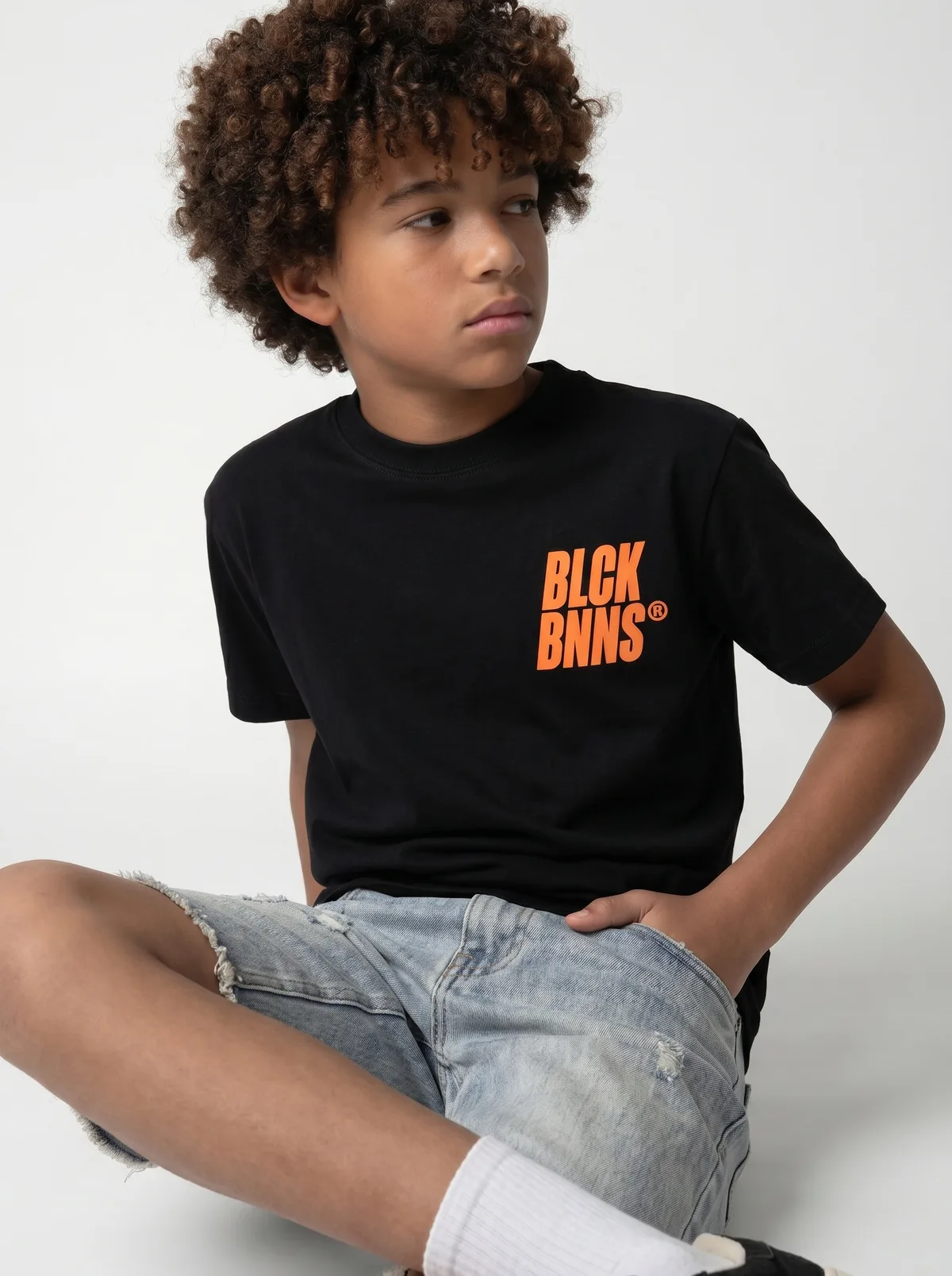 Black Bananas black BLCK BNNS orange-print tee with light denim shorts editorial side-walking pose on AI young model in clean studio