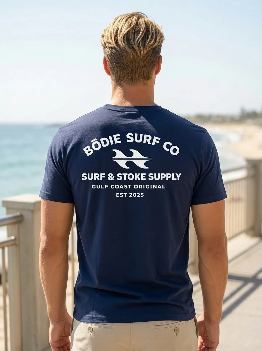Bōdie Surf Co navy tee back graphic showing full Surf and Stoke Supply logo, AI model facing ocean from boardwalk
