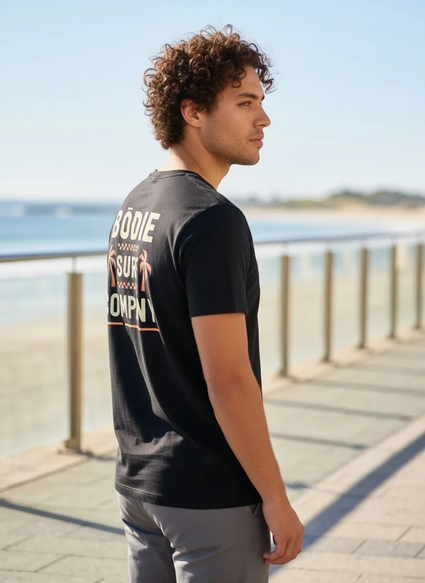 Bōdie Surf Co black tee with palm tree back graphic on AI male model on sunlit boardwalk with ocean horizon