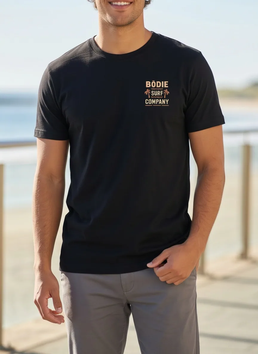Bōdie Surf Co black tee with small chest logo on AI male model facing camera on sunlit boardwalk with ocean railing