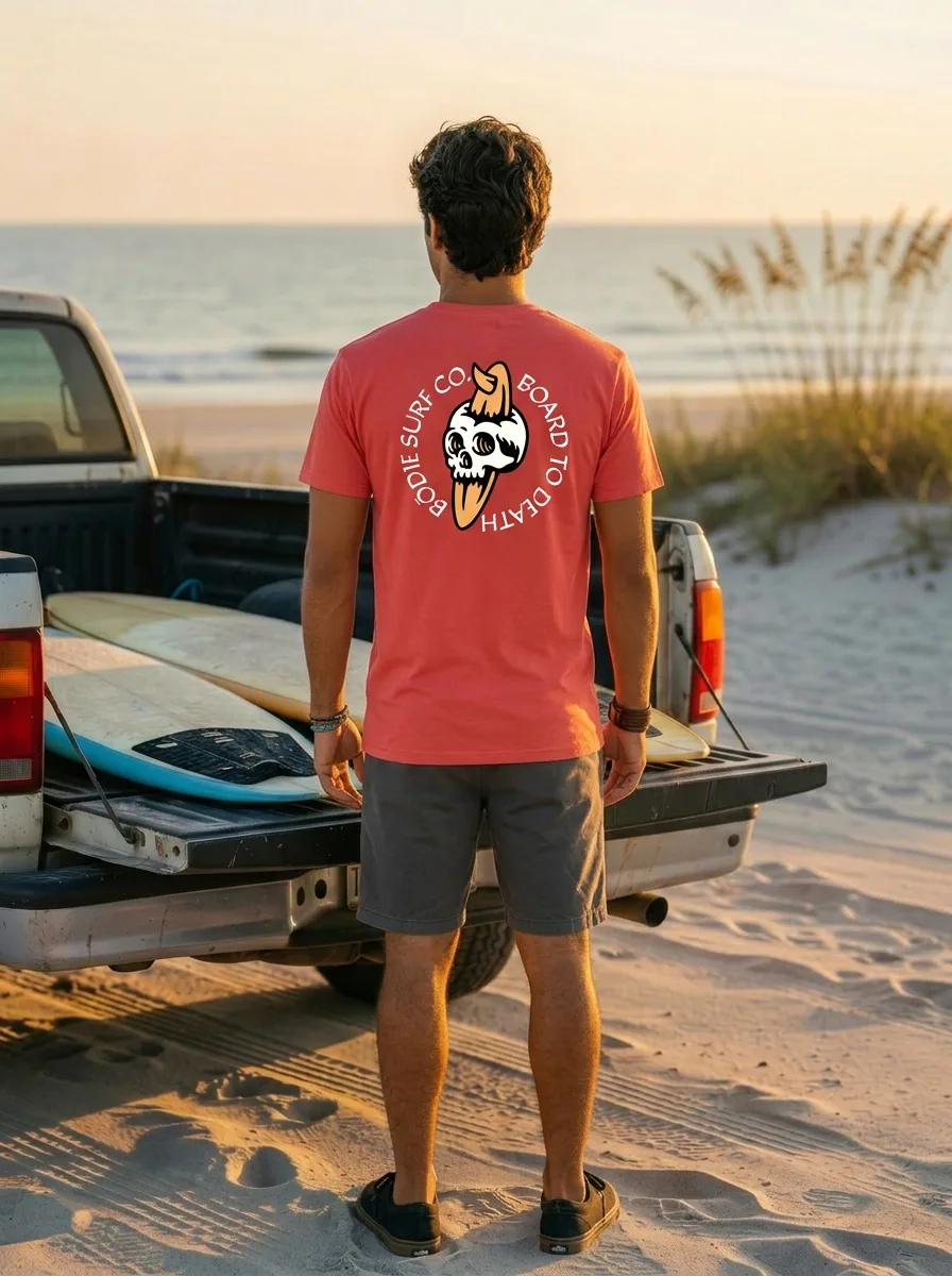 Bōdie Surf Co coral tee with BOARD TO DEATH skull-and-surfboard back graphic on AI male model loading boards into pickup truck at sunset