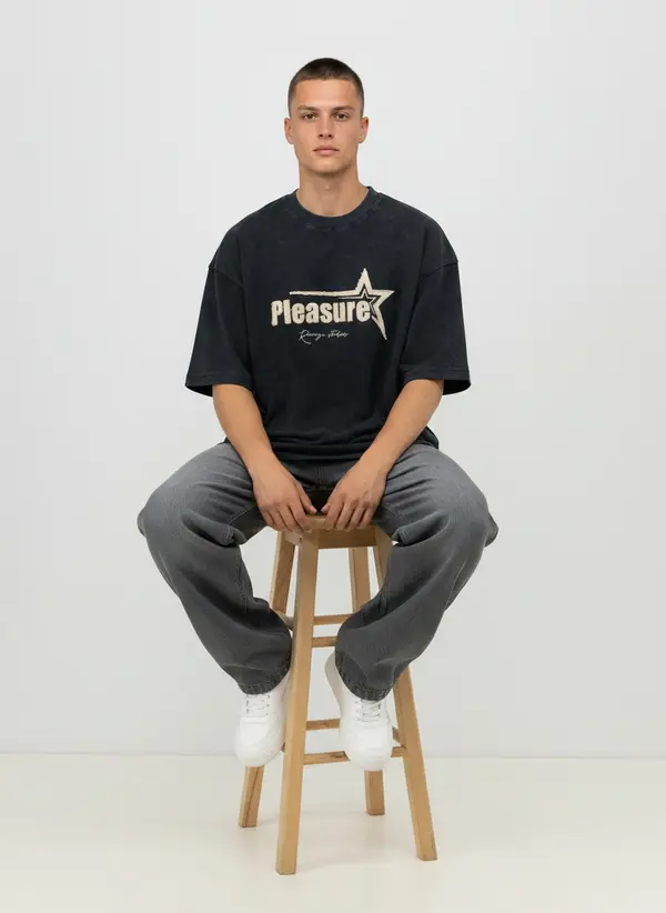 Bully Pleasure washed black oversized tee, seated on a stool with grey denim