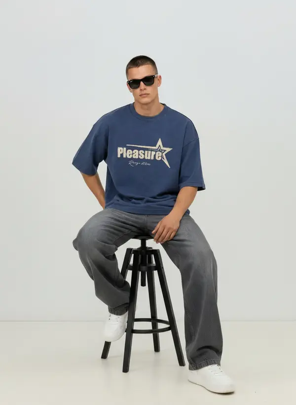 Bully Pleasure navy oversized tee, seated on a stool with grey denim