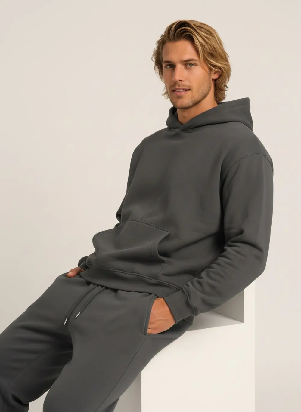 Future Homme graphite hoodie and jogger set on an AI male model seated on a block, hood down, warm bone studio