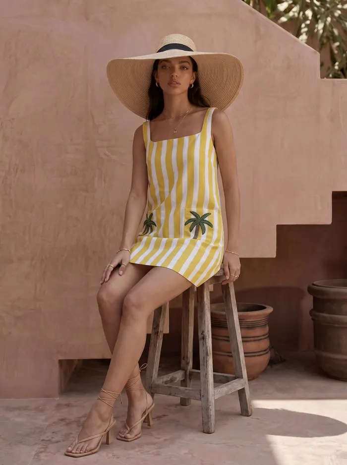 Maison Blu yellow stripe palm-pocket mini dress with wide-brim straw hat seated editorial pose on rustic wooden stool against Marrakech terracotta wall and clay pot