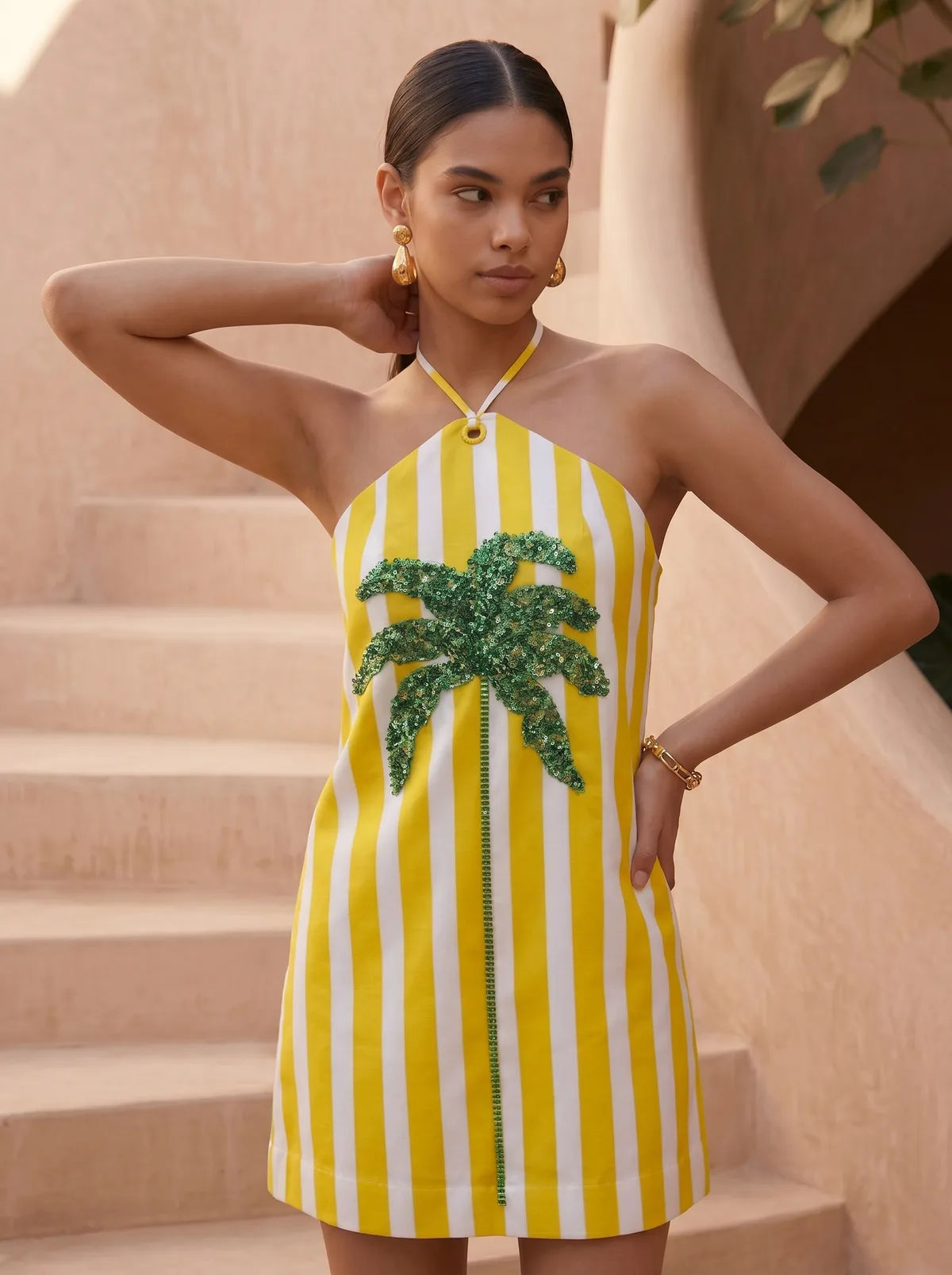 Maison Blu yellow stripe sequin-palm halter mini dress editorial three-quarter close-up on AI model with gold drop earrings against terracotta stairwell