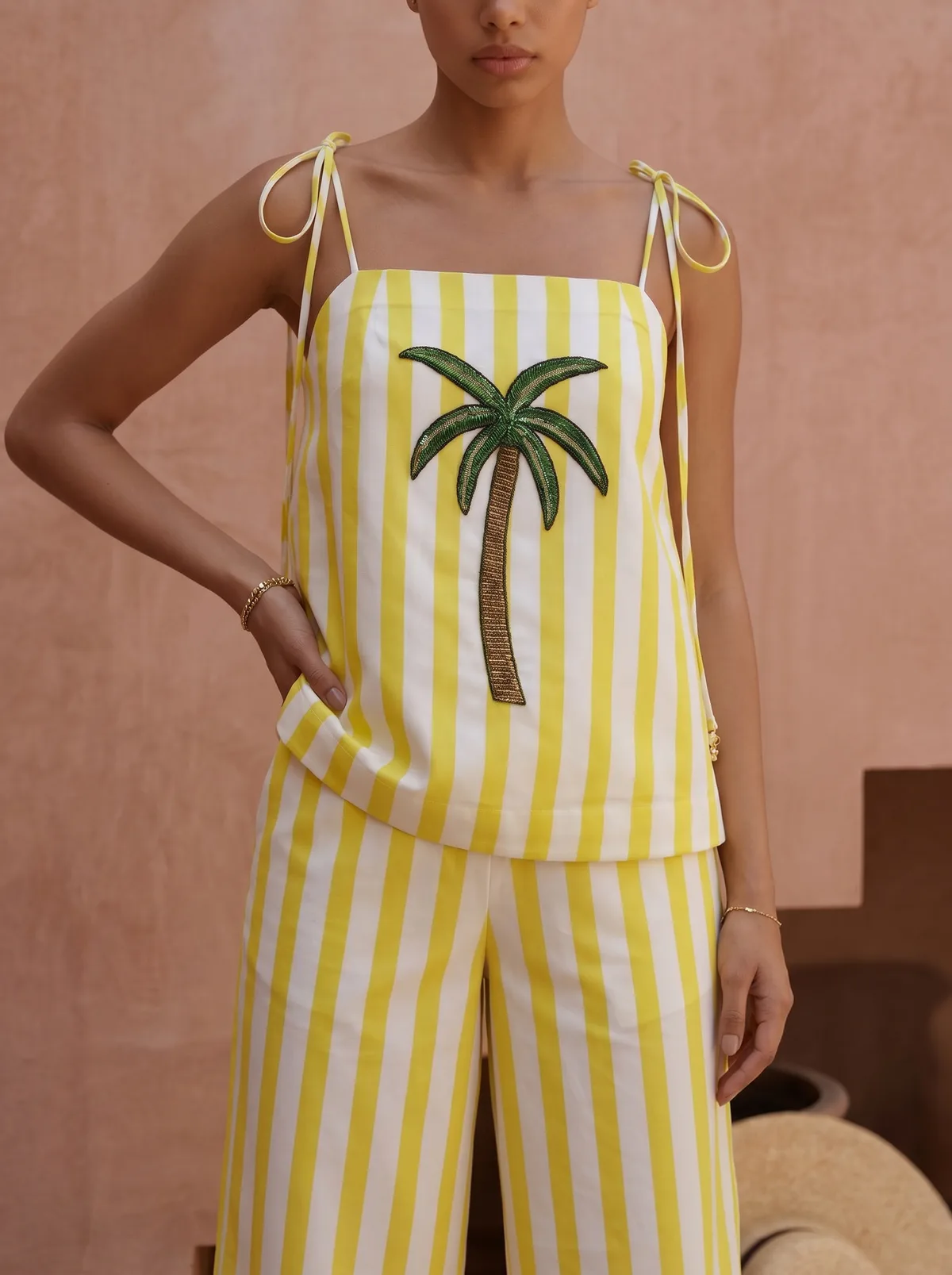 Maison Blu yellow stripe palm halter tank close-up showing tied-bow shoulder straps and large green sequin palm-tree motif against pink-stucco wall