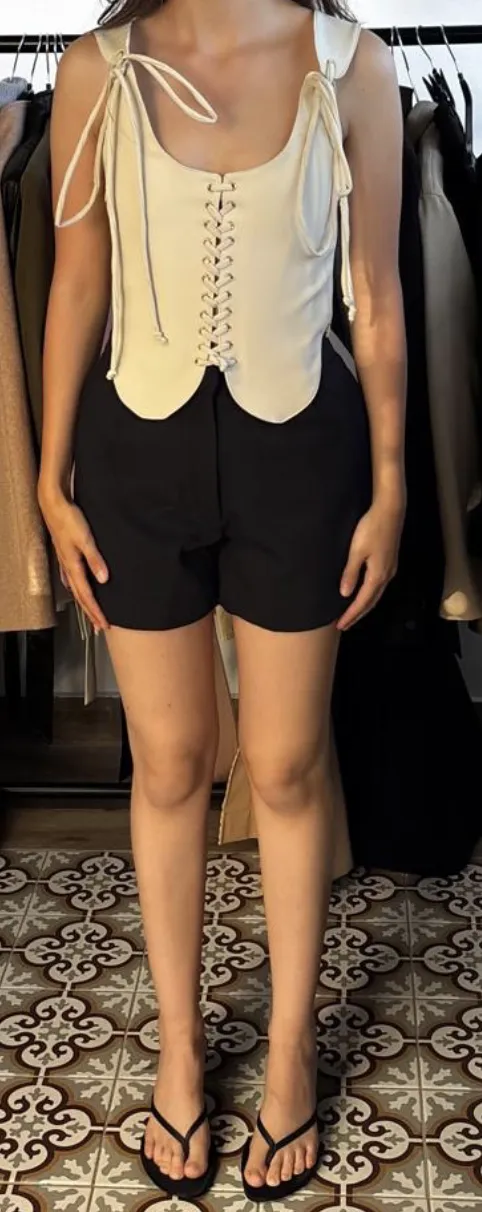 Input showroom photo — the lace-up vest top in cream on a fit model in the showroom, phone shot, tiled floor and clothing rail behind