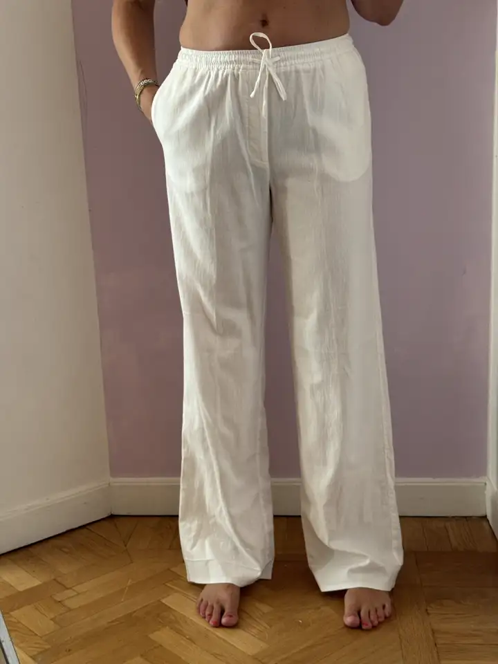 Seller's apartment selfie wearing cream-colored linen pants against a purple wall with wood parquet floor and an ironing board visible in the background, the original input for the MODA AI catalog generation