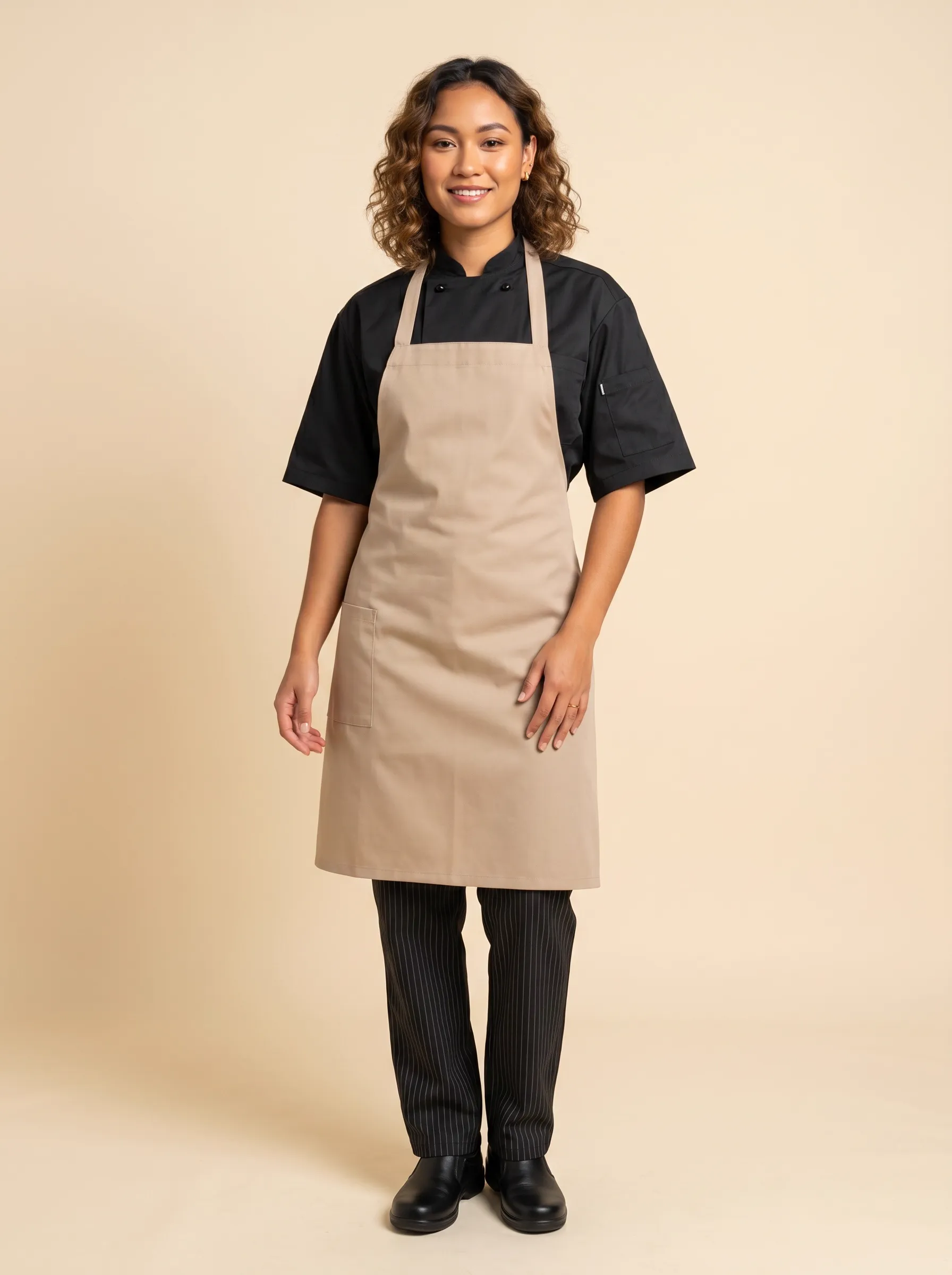 MODA AI output — female chef in tan bib apron over black short-sleeve chef coat with black-and-white striped chef pants, three-quarter front pose on warm beige studio backdrop, demonstrating in-catalog imagery for chef uniform brands