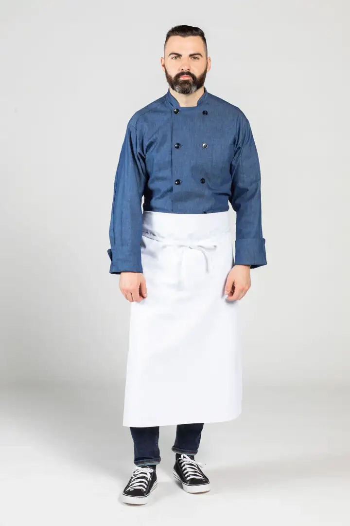 Input — bearded male chef in denim long-sleeve chef coat with white waist apron, jeans, and Converse sneakers on plain studio background, the original supplier catalog photograph for the casual chef uniform set