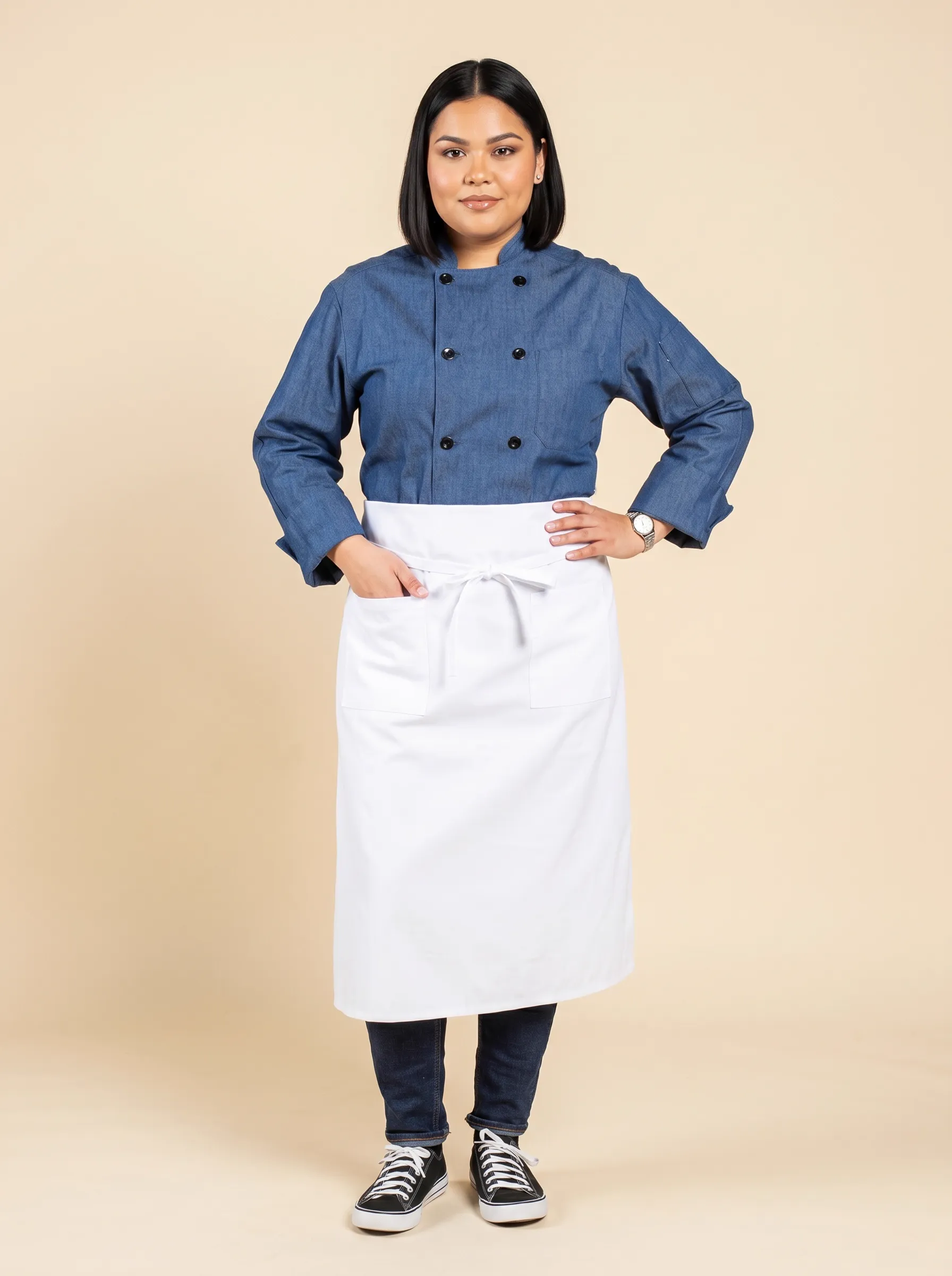 MODA AI output — plus-size Latina female chef in denim chef coat, white waist apron, dark jeans and black Converse sneakers, three-quarter front pose with hand on hip on warm beige studio backdrop, demonstrating body diversity for chef uniform catalogs
