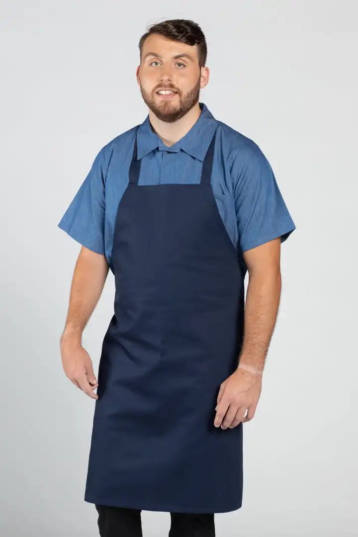 Input — male model in navy bib apron over chambray short-sleeve shirt with jeans and white sneakers on plain studio background, the original supplier catalog photograph for the navy bib apron set