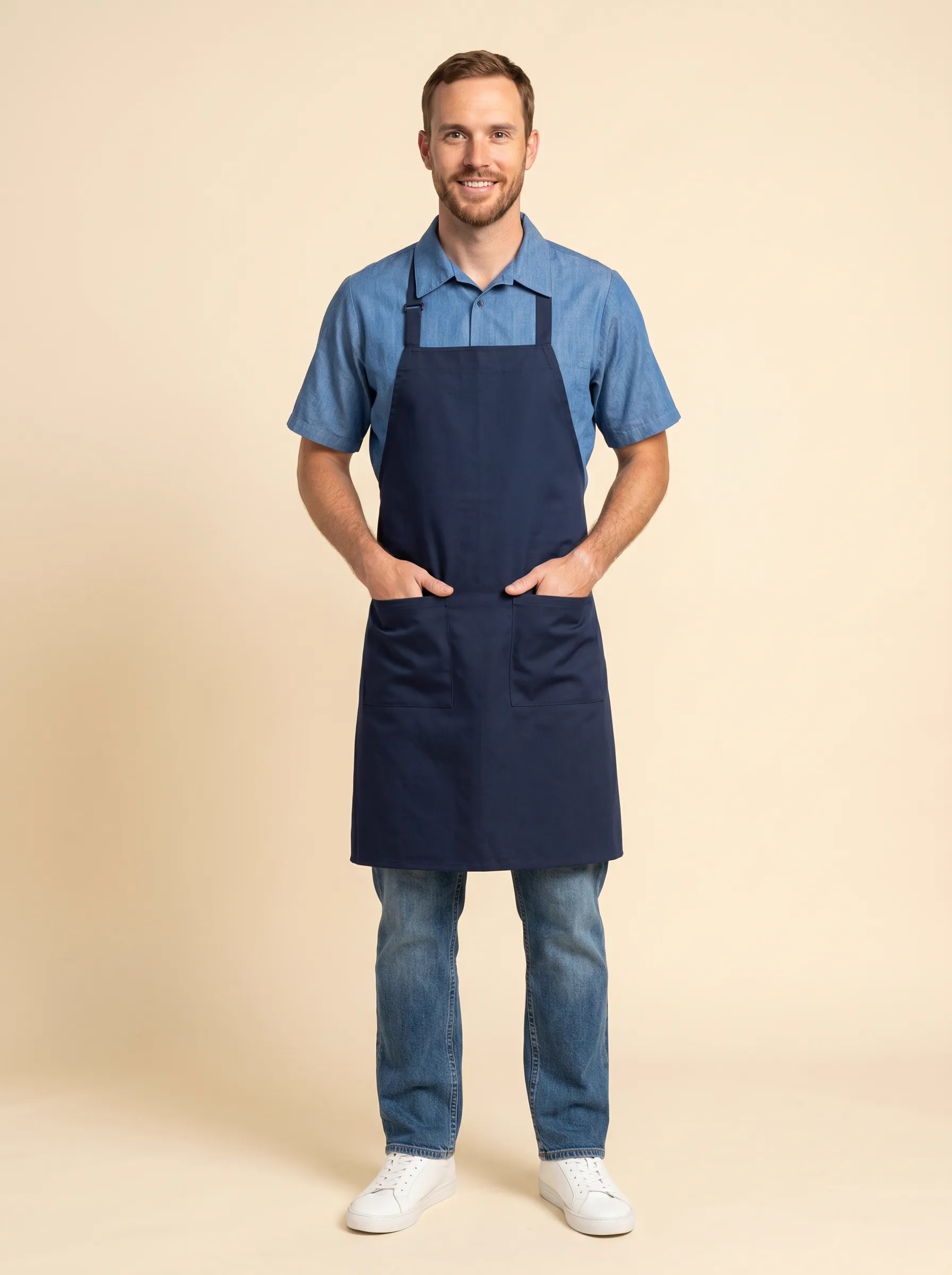 MODA AI output — bearded male server in navy bib apron over chambray short-sleeve shirt, jeans and white sneakers, front pose with hands in apron pockets on warm beige backdrop, demonstrating front-of-house uniform catalog imagery