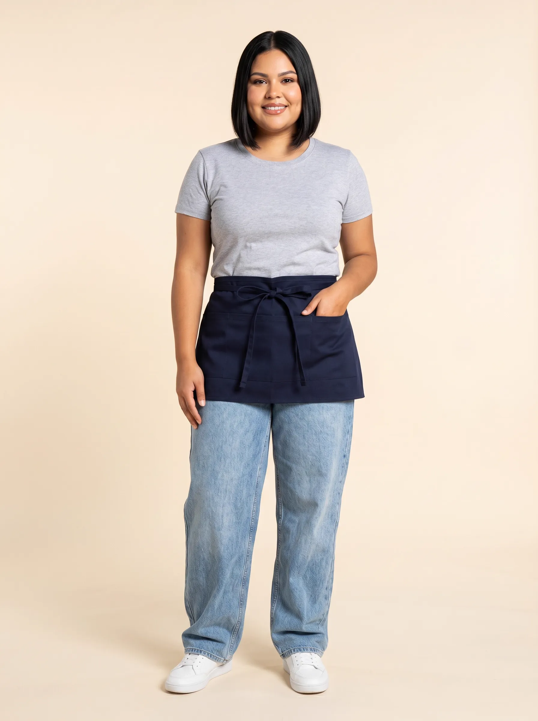 MODA AI output — plus-size Latina female server in short navy waist apron, gray tee and light wash jeans with white sneakers, front pose on warm beige backdrop, demonstrating server uniform catalog imagery on a real body