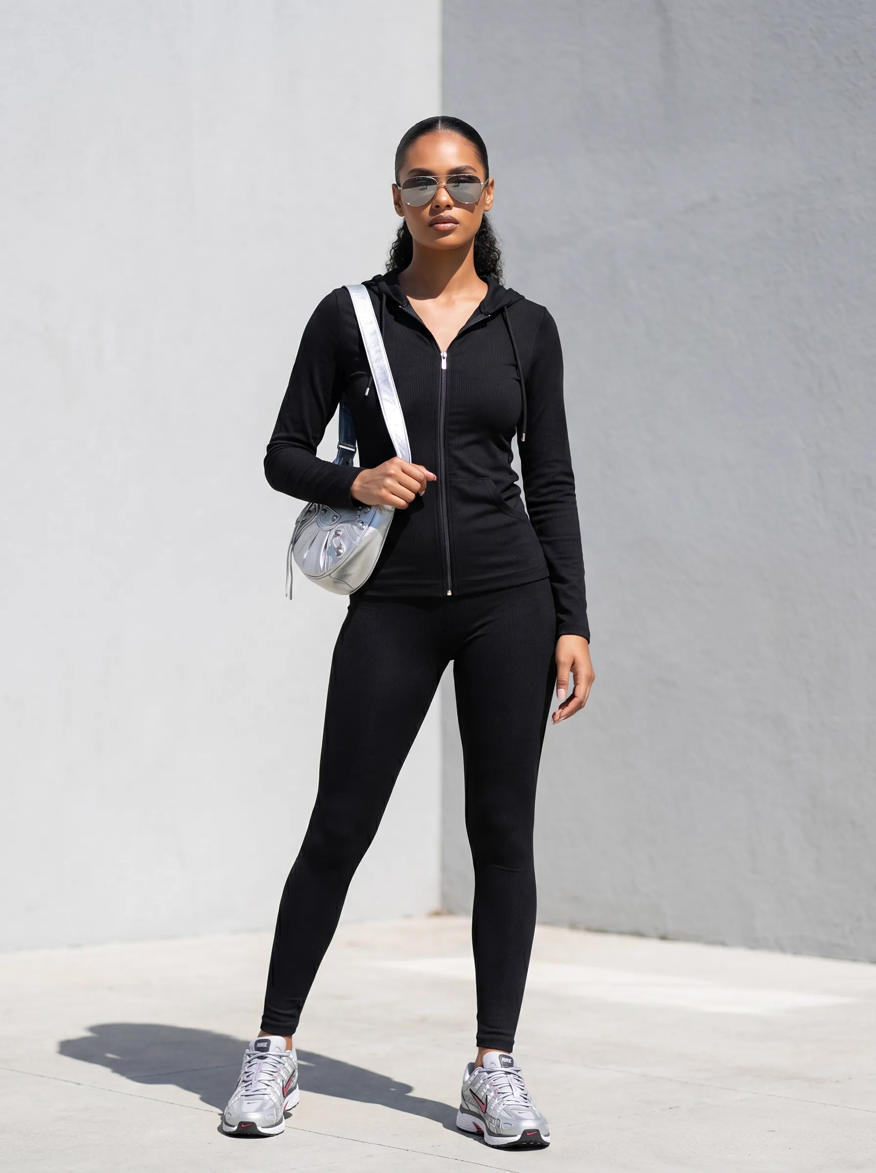 MODA AI recataloging hero — Black model in black athleisure hoodie and leggings styled with silver crossbody bag, aviator sunglasses, and silver running sneakers against sunlit urban concrete wall, demonstrating seasonal product photography refresh for Shopify boutiques
