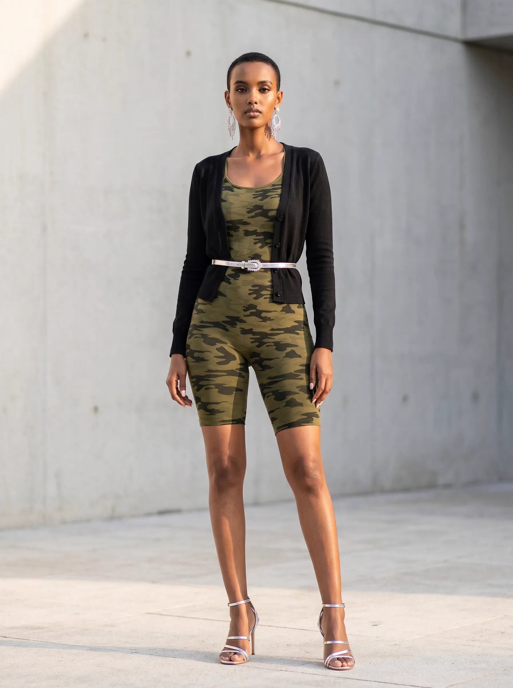MODA AI recataloging output — buzz-cut Black model in same camo romper styled with cropped black cardigan, silver belt, silver strappy heels, against sunlit concrete urban wall, elevated streetwear restyle