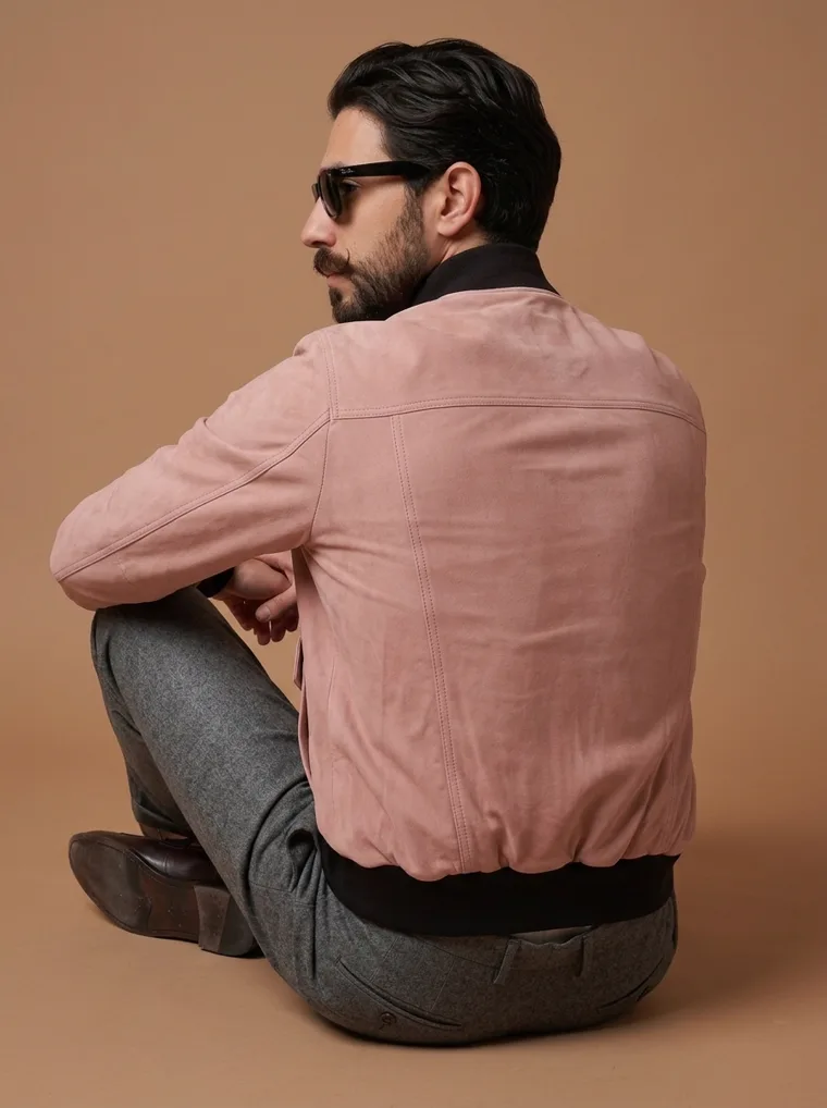 A dusty blush bomber jacket worn with grey wool trousers and a black roll neck against a warm caramel backdrop. FW26 look 01, Relaxed Bomber in Blush, frame 5 of 5