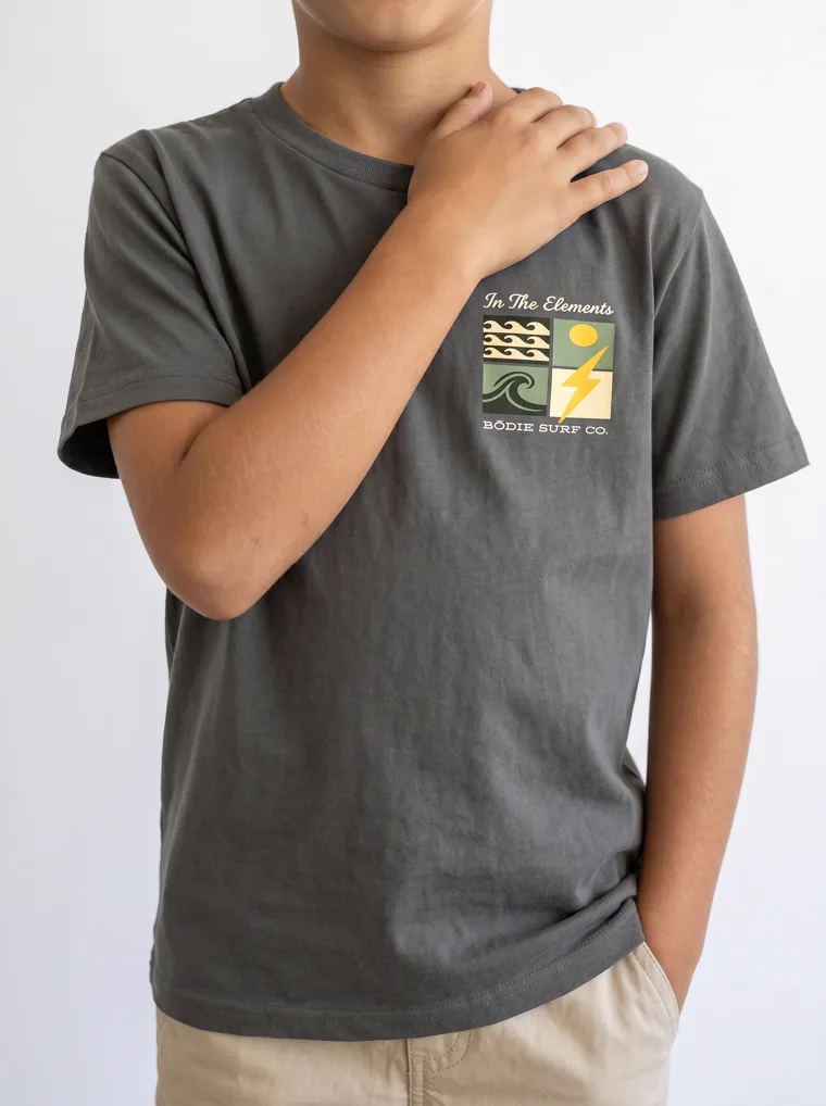 A charcoal boys tee with a surf graphic on the chest and a larger version across the back, worn with stone shorts. FW26 look 18, Graphic Basic in Charcoal, frame 2 of 5