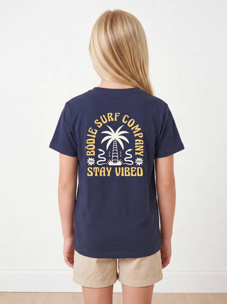 The back of a navy girls tee showing a palm and surfboard graphic, FW26 look 19