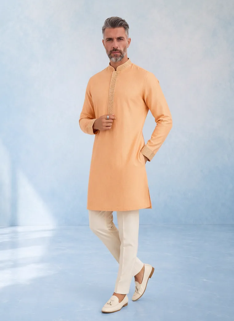 A model in a peach kurta with cream churidar trousers and cream loafers, photographed against a pale blue painted backdrop, FW26 look 03