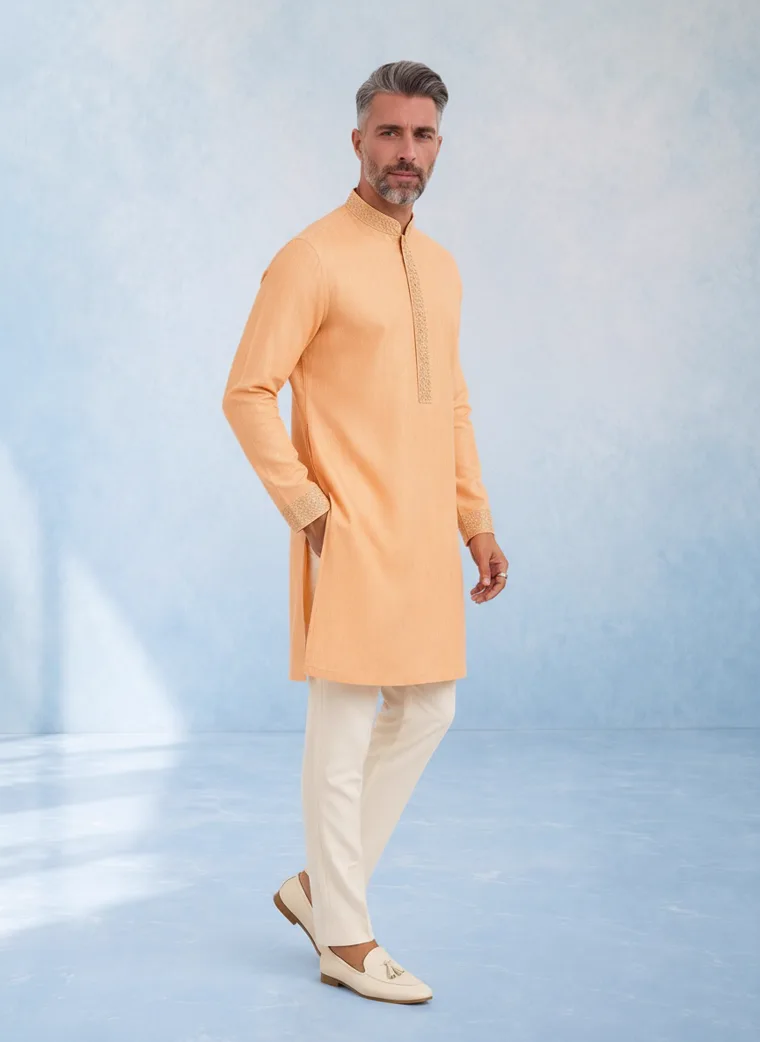 A peach kurta with cream churidar trousers and cream loafers against a pale blue painted backdrop. FW26 look 03, New Neutral in Peach, frame 2 of 5
