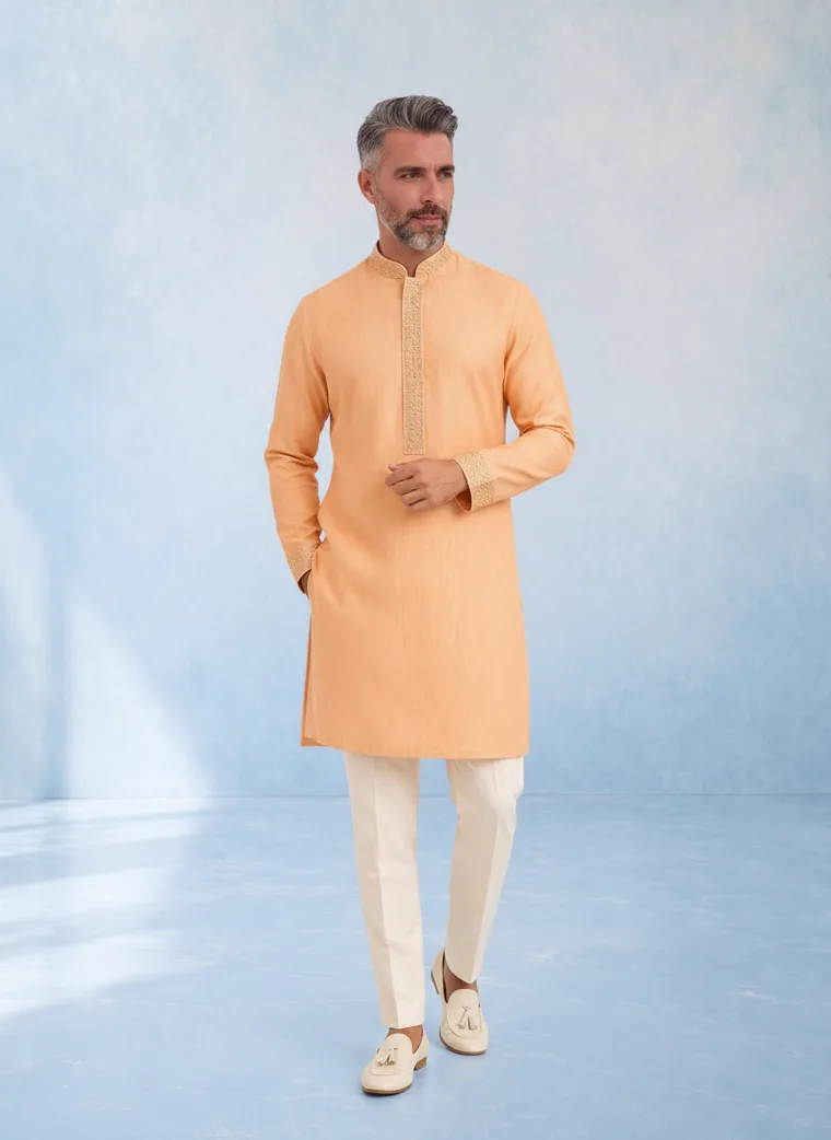 A peach kurta with cream churidar trousers and cream loafers against a pale blue painted backdrop. FW26 look 03, New Neutral in Peach, frame 5 of 5