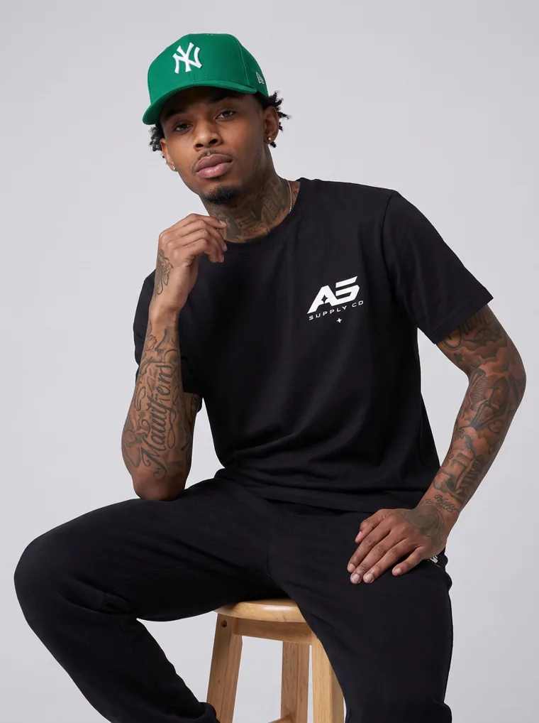 A model in a black tee and black trousers with a green baseball cap, FW26 look 07
