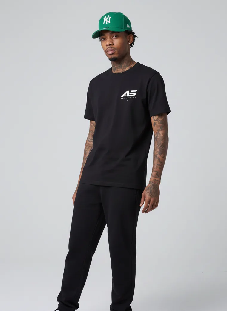 A black tee and black trousers worn with a green baseball cap and a small chest logo. FW26 look 07, Street Uniform in Black, frame 2 of 5