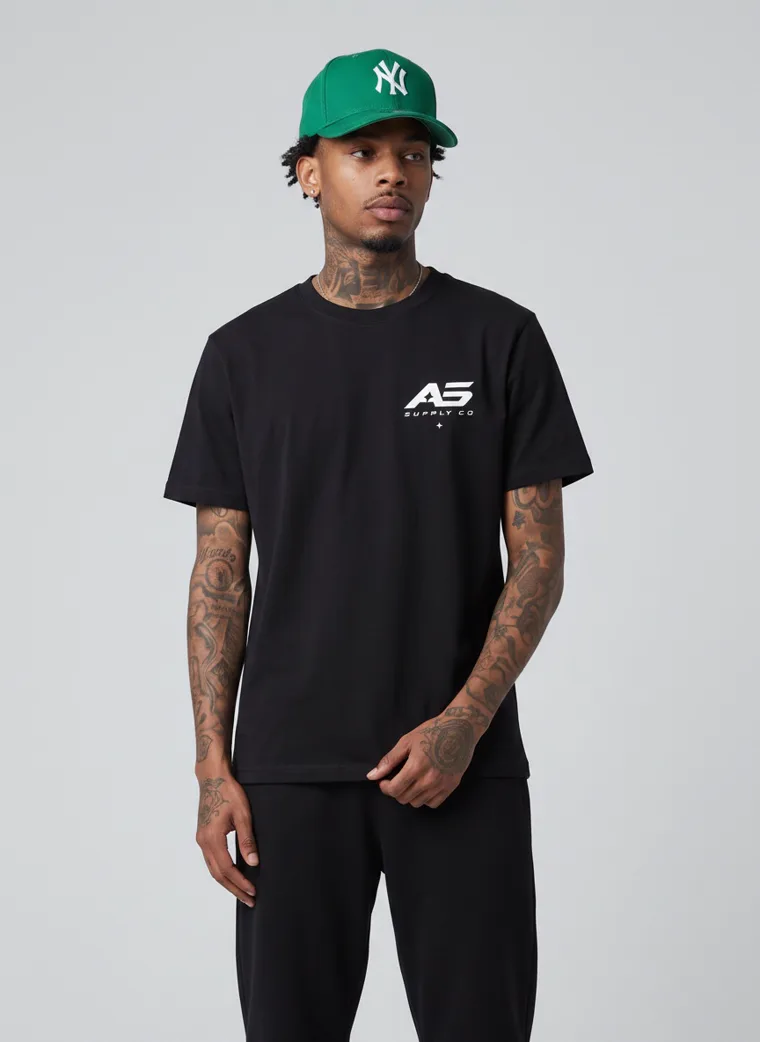 A black tee and black trousers worn with a green baseball cap and a small chest logo. FW26 look 07, Street Uniform in Black, frame 3 of 5