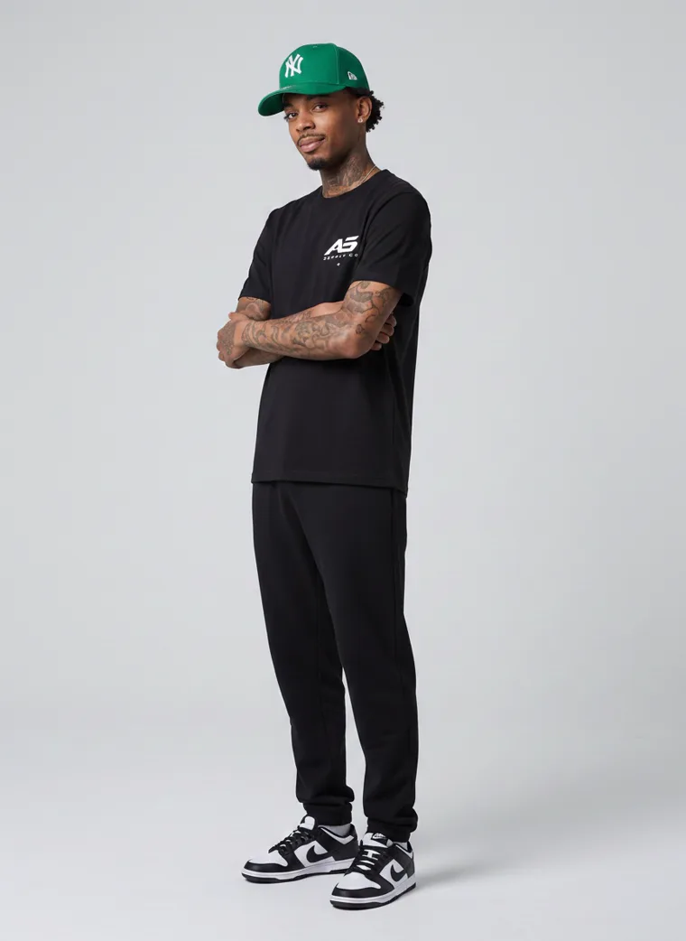 A black tee and black trousers worn with a green baseball cap and a small chest logo. FW26 look 07, Street Uniform in Black, frame 4 of 5
