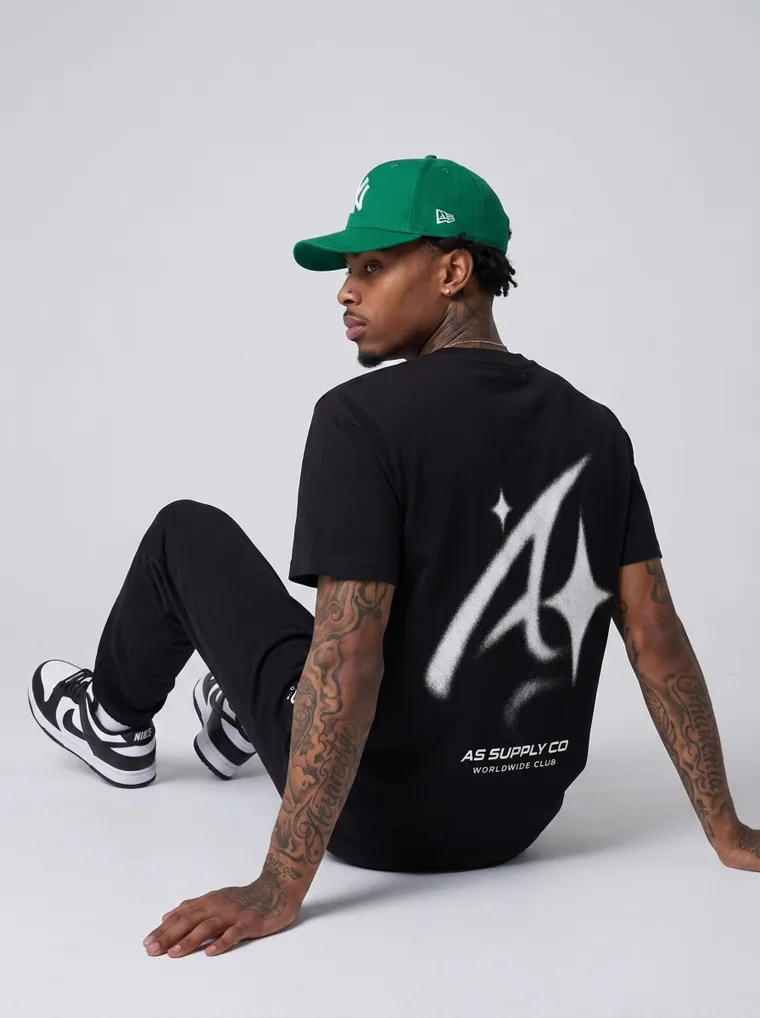 A black tee and black trousers worn with a green baseball cap and a small chest logo. FW26 look 07, Street Uniform in Black, frame 5 of 5