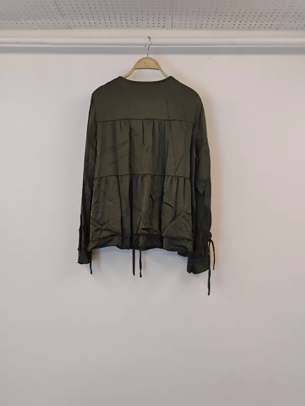 Hanger shot input — the back of the same olive tiered blouse, turned on the hanger against the shop wall