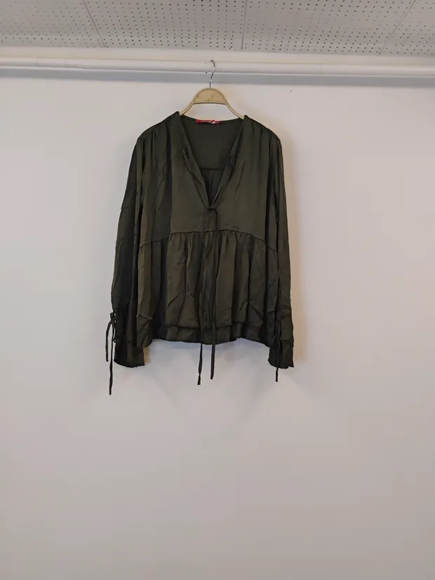 Hanger shot input — an olive green tiered blouse with tie cuffs hanging on a wooden hanger against a plain shop wall