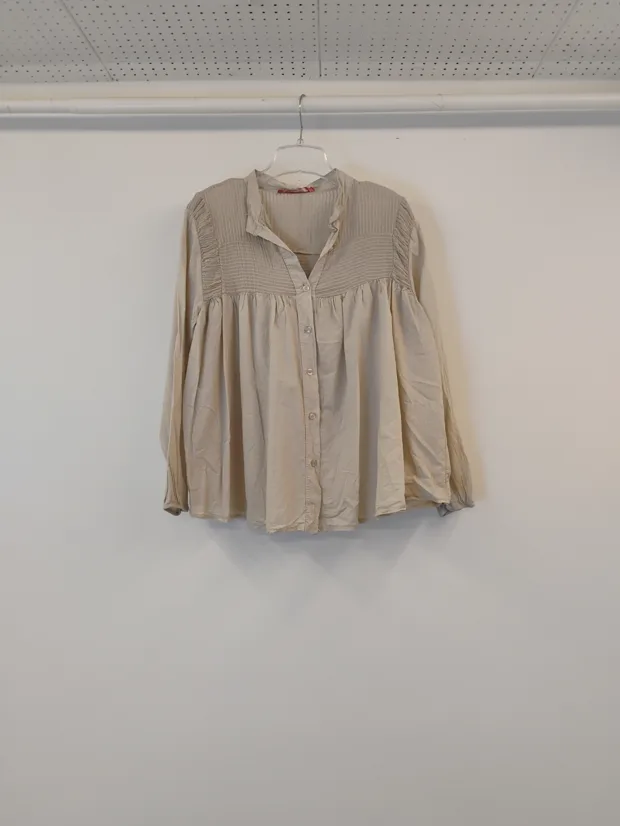 Hanger shot input — an oatmeal smock blouse with a gathered yoke, hanging on a hanger against a plain shop wall