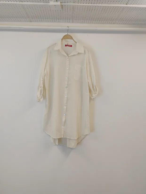 Hanger shot input — a long cream button-through shirt dress with a curved hem, hanging against a plain shop wall