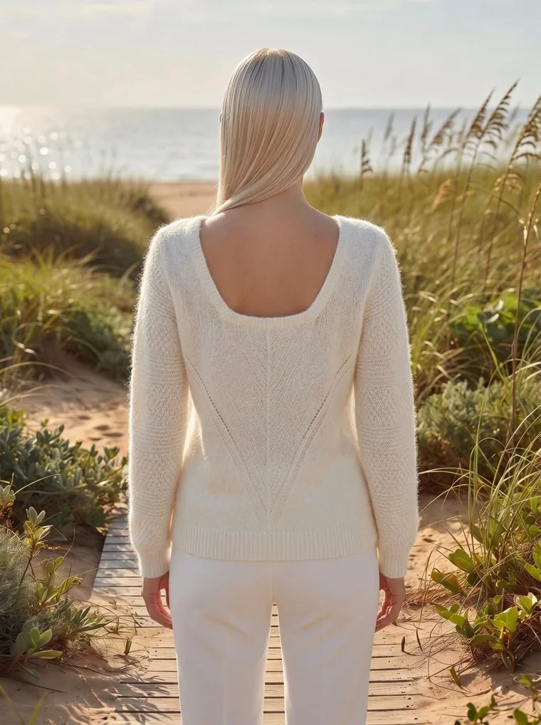 A cream v-neck knit with white trousers on a wooden boardwalk through dune grass by the sea, in late sun. V-Neck Knit in cream, back view