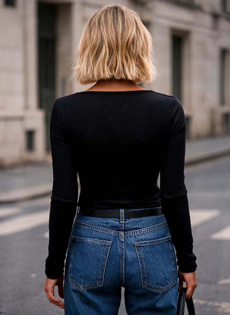 The same black scoop-neck long-sleeve top with straight blue jeans, black loafers and a small black bag, on a city crosswalk in low sun. Scoop-Neck Long-Sleeve Top in black, back view, close