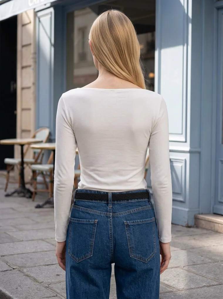 An ivory ribbed henley top with straight dark jeans and white trainers, in front of a pale blue cafe facade with bistro chairs. Ribbed Henley Top in ivory, back view
