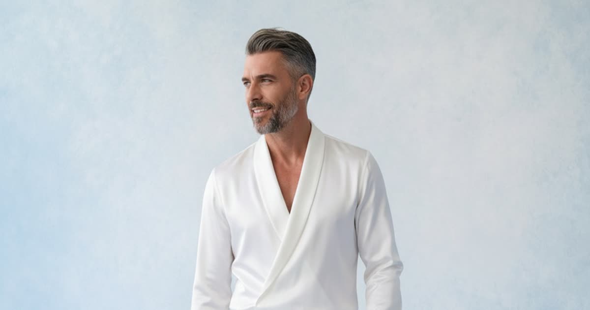 A man in an all white shawl collar shirt and trousers photographed against a soft blue backdrop, a pale garment kept legible by a coloured ground