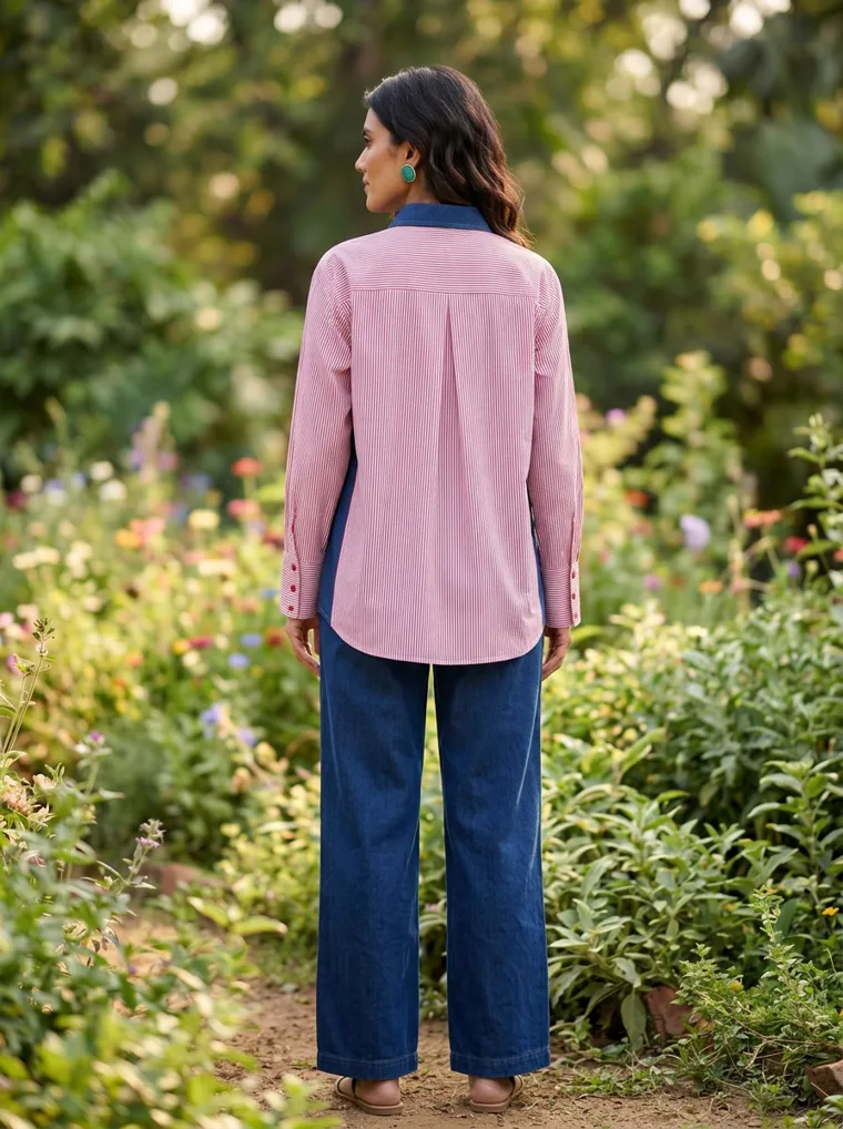 A pink shirt with a large floral embroidery and navy trim, worn with blue wide leg trousers in a flower garden. Embroidered Shirt in Pink, frame 5 of 5
