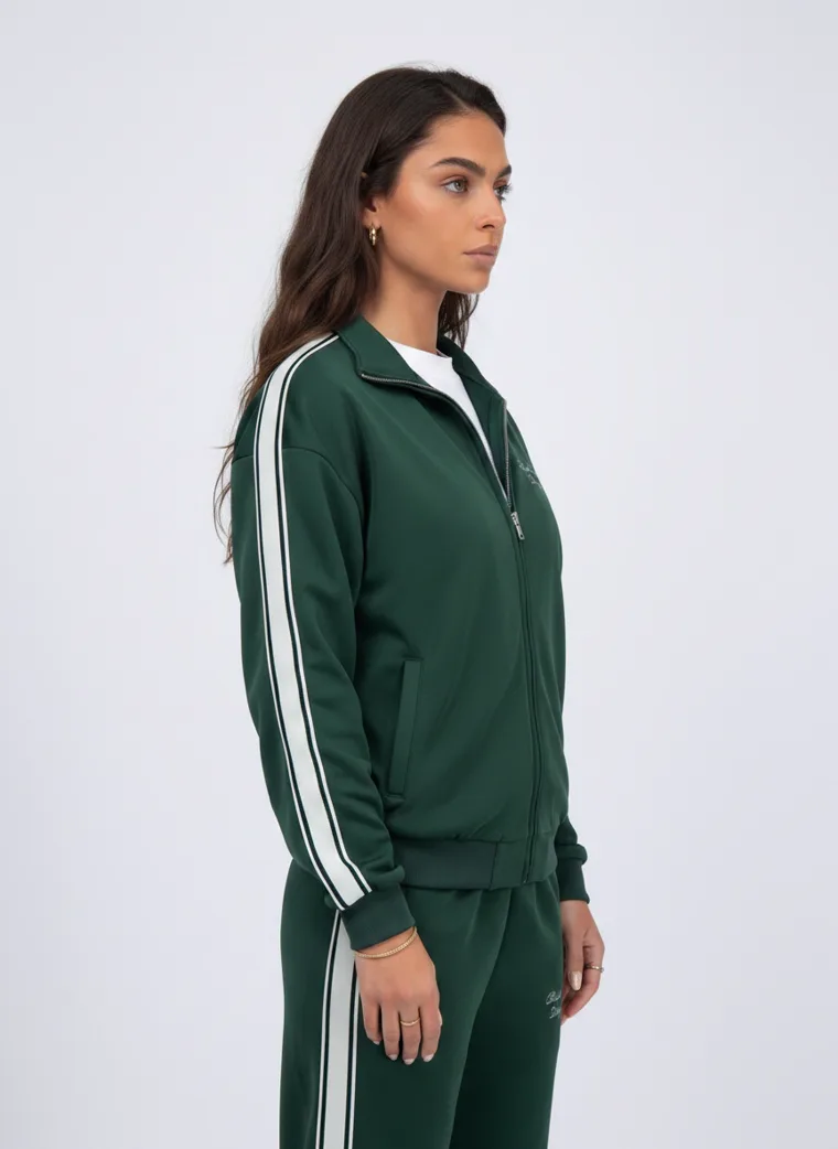 A forest green track jacket and pants with white side stripes and small embroidered chest text on pale grey. Track Jacket and Pants in Forest, frame 4 of 5