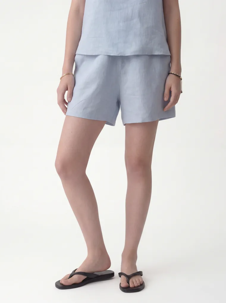 A pale blue linen sleeveless top with matching linen shorts and black flip flops on white. Linen Top and Shorts in Pale Blue, frame 4 of 5