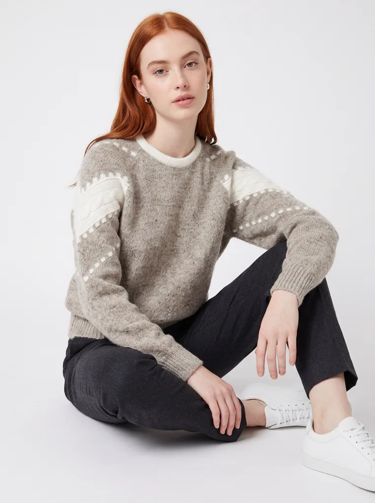 A grey marled crew-neck knit with white intarsia stripes and dots down the sleeves and a cream neckline, worn with black trousers. Intarsia Stripe Knit in grey, seated