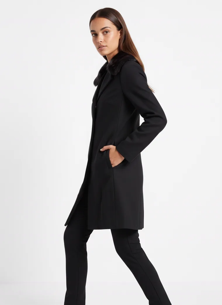 A model in a black wool coat with a faux-fur collar, full length in three-quarter, the coat falling from the shoulder to below the knee