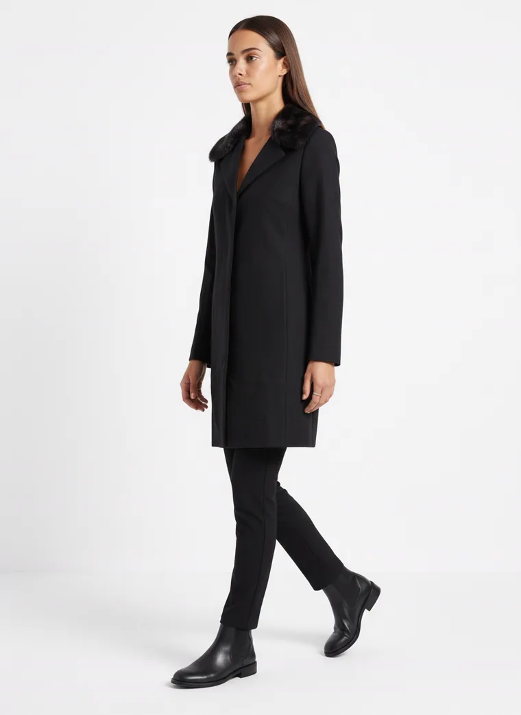 A model walking in a black wool coat with a faux-fur collar, the hem swinging behind her