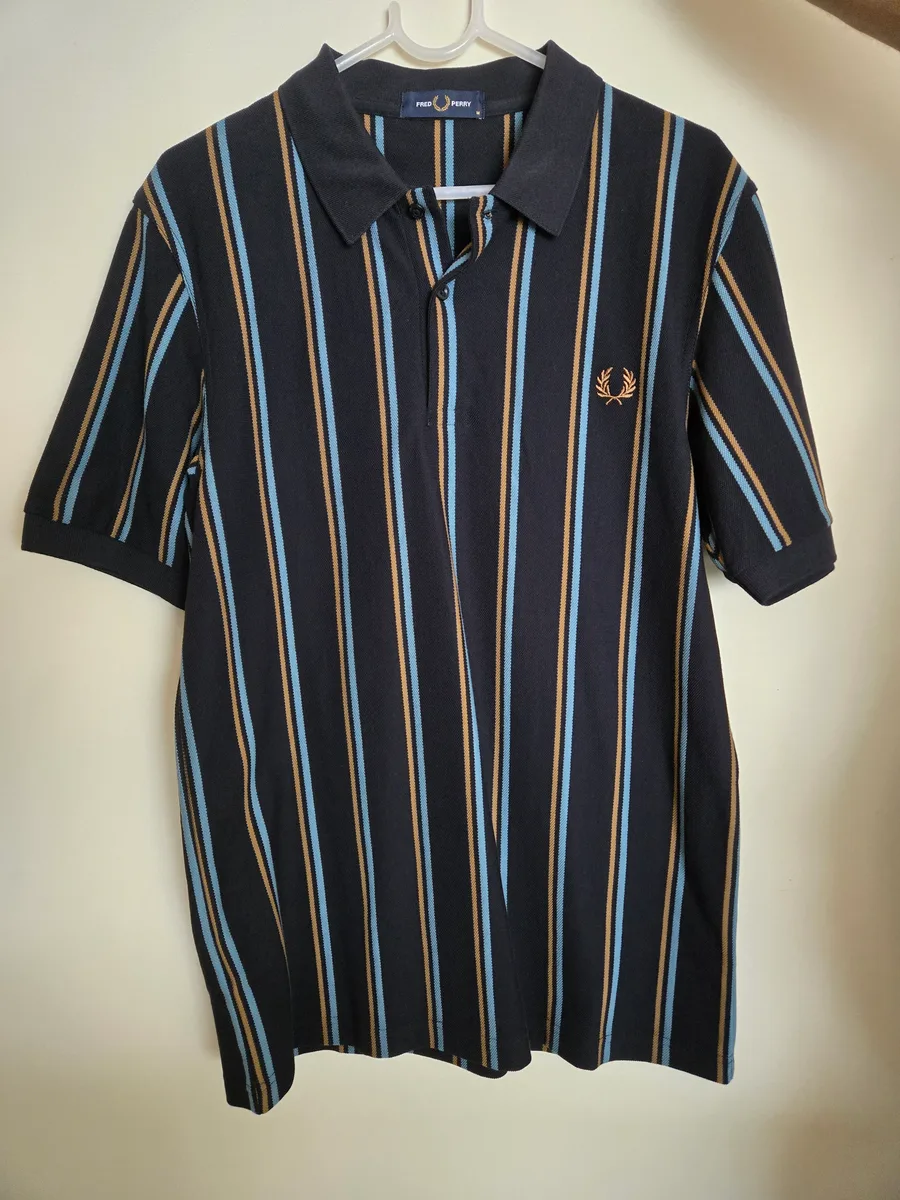 Input hanger shot, front. A navy pique polo with tan and pale blue vertical stripes on a wooden hanger against a plain wall, with an embroidered laurel wreath logo on the chest and a buttoned placket
