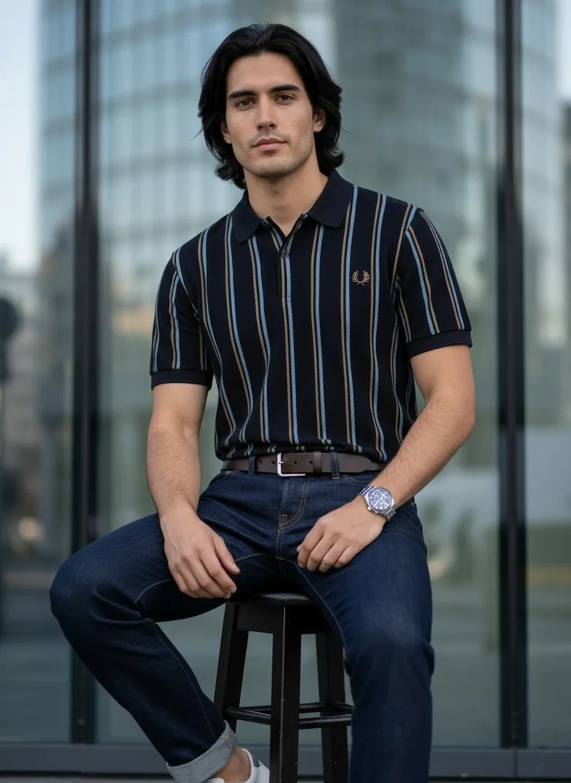 MODA AI shot 5, the same model seated on a stool in the striped navy polo with dark jeans, a brown belt and a watch, in the city setting